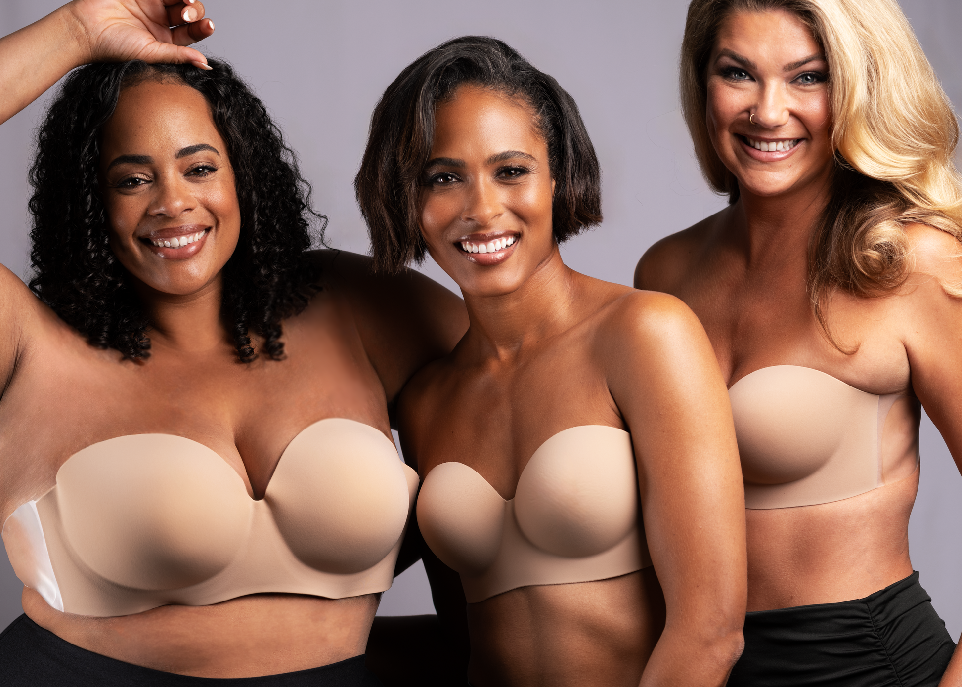 3 models of different sizes stand together smiling, wearing the Voluptuous Backless Strapless Bra together, showing the product range and how it lifts and shapes. Bra is nude in color.