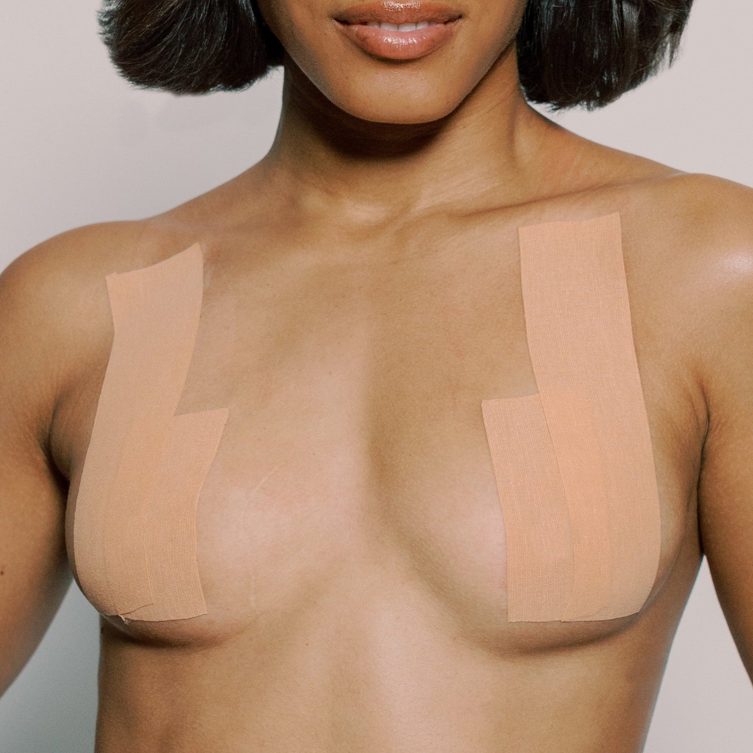 Model wears a few strips of tape from the Tape It Your Way breast tape roll. They are placed vertically showing how the tape can lift and shape. The strips are spaced, leaving plenty of space in the center of the chest for plunging necklines. Tape is nude in color.